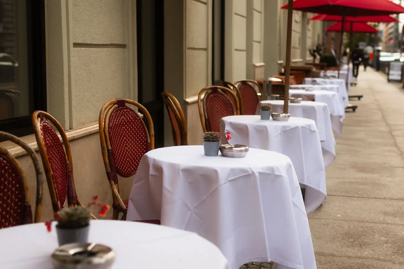 A row of sidewalk tables with white cloths and red umbrellas outside a restaurant