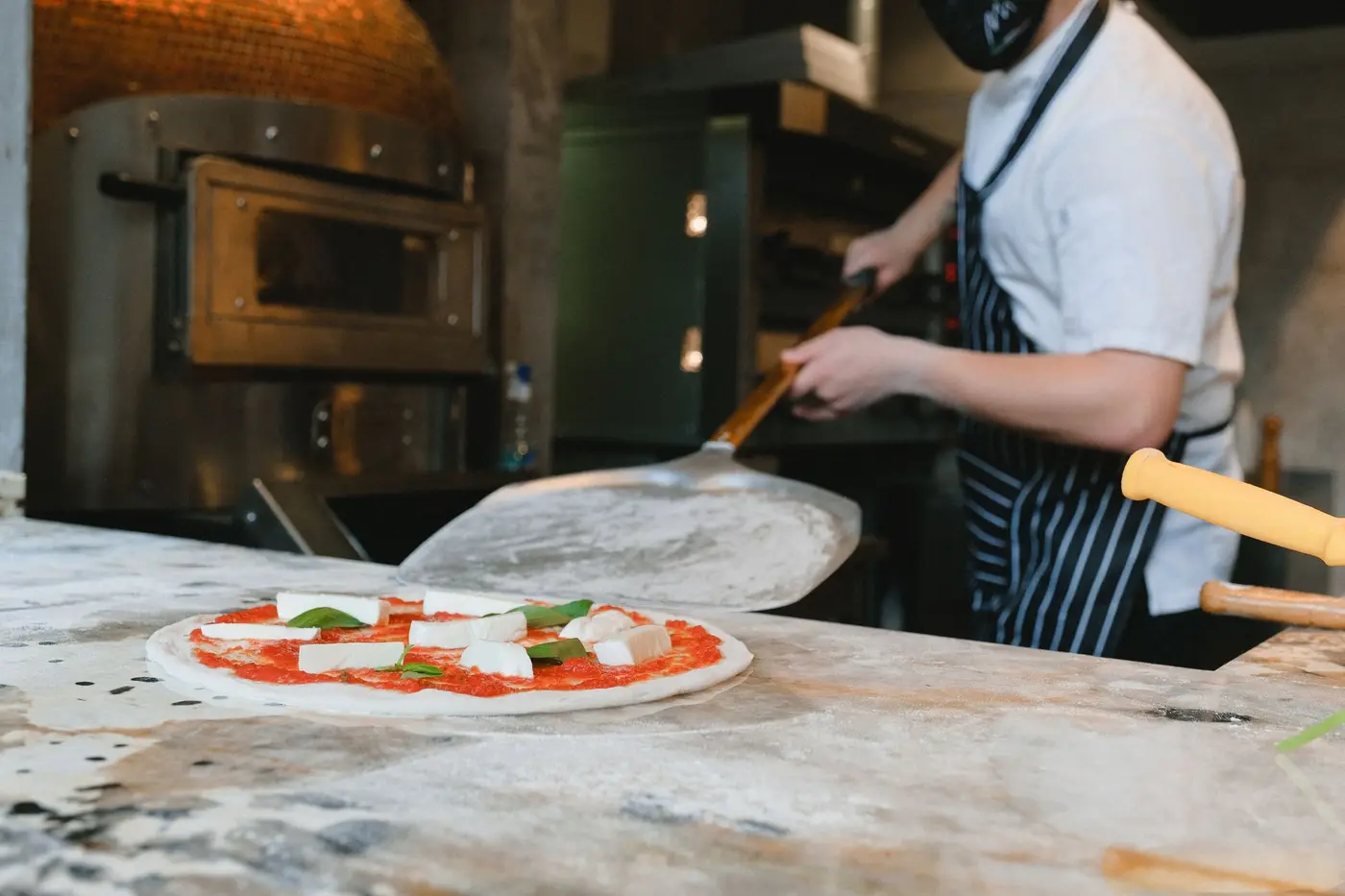 A pizza on a peel beside a pizzeria oven